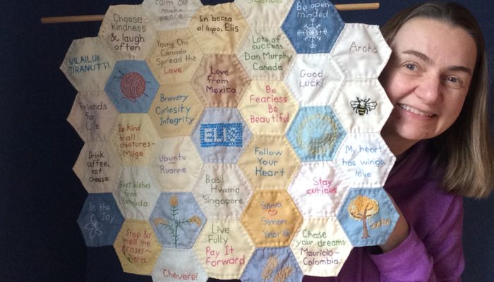 Quilter holds up small quilt made from hexagon-shaped blocks with messages of encouragement sewn on each block