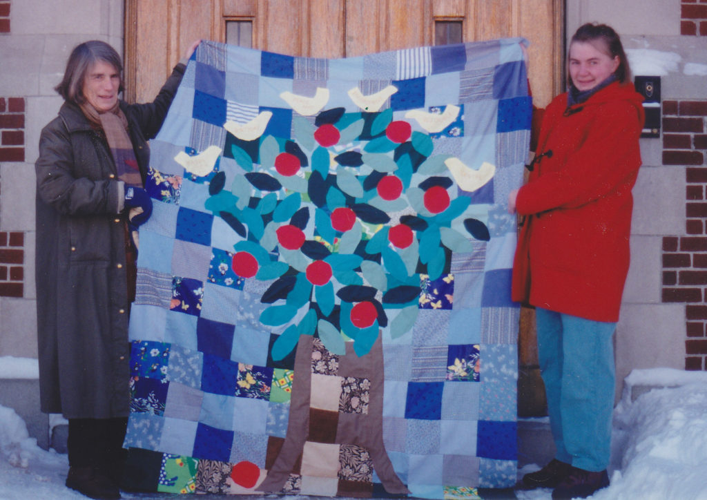 Two women hold up a quilt showing a tree with leaves, apples and birds on it made from scrap fabrics