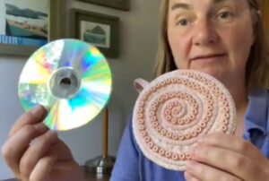 Artist holding up a swirly peach ornament beside a recycled CD to show how it fits inside