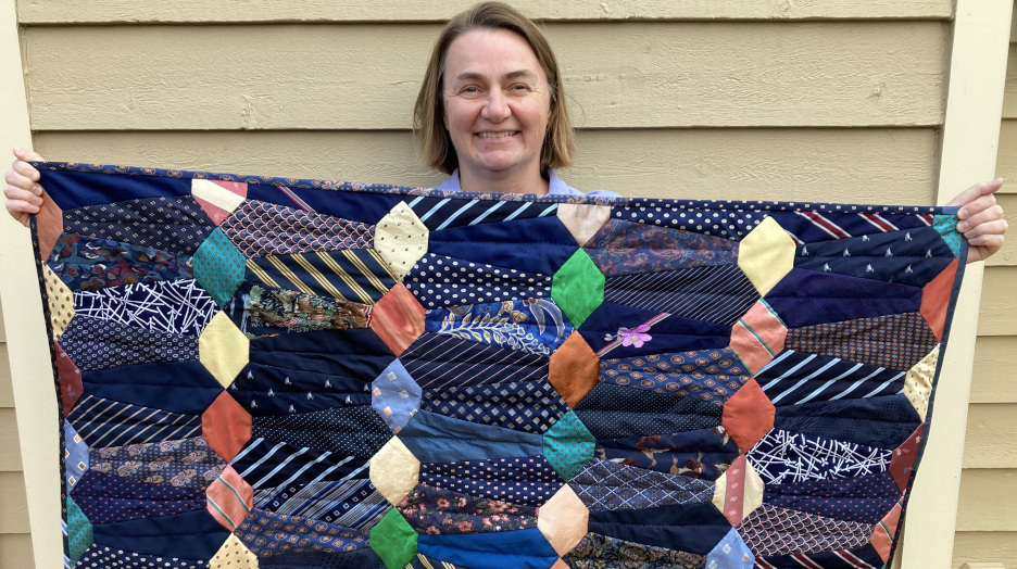 Woman holds up a quilt that is made from mostly navy blue neckties with some colorful diamond shape parts as well