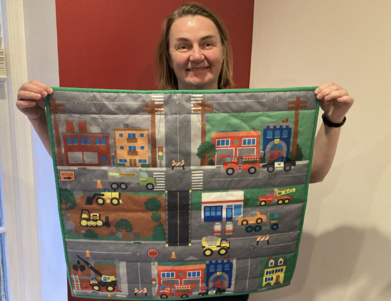 A woman holds up a colorful fabric panel with many images of trucks and streets - a city scape