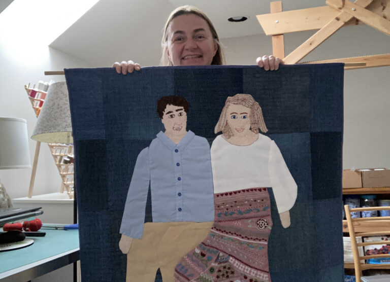 A woman holds up a quilt with a denim blue background. The quilt shows embroidered portraits of two people sitting.