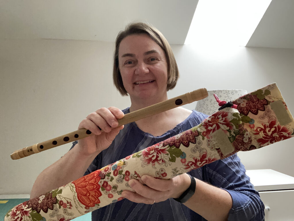 A woman holds up a bamboo flute in one hand and a long tube-shaped case covered with beautiful Japanese fabrics with a base color of beige, decorated with a design of pink and orange flowers